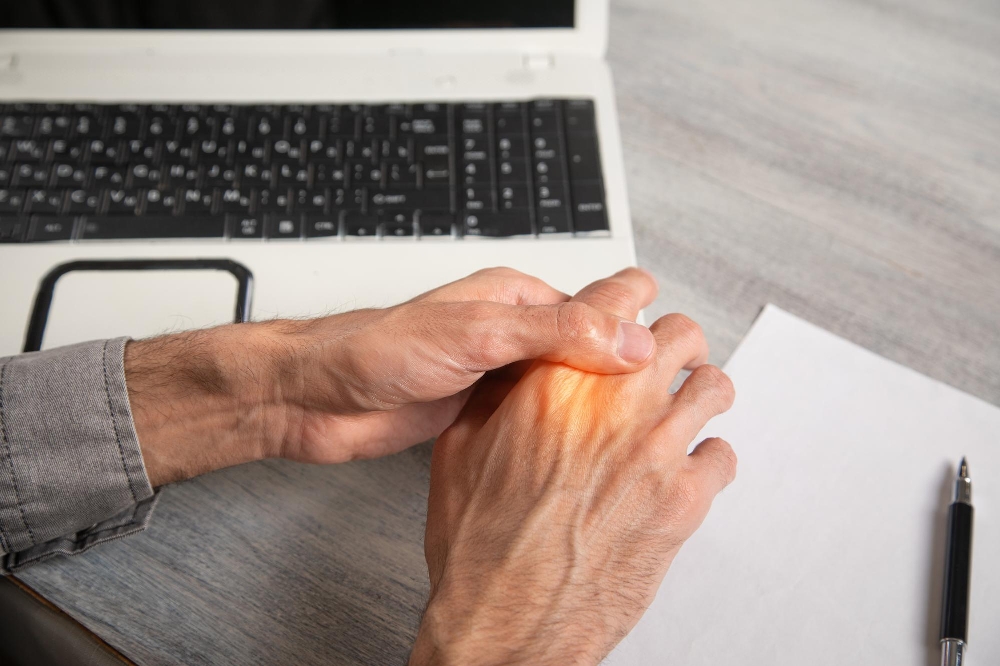 Man at a laptop pressing his thumb into the back of his other hand, an everyday setting for joint protection techniques